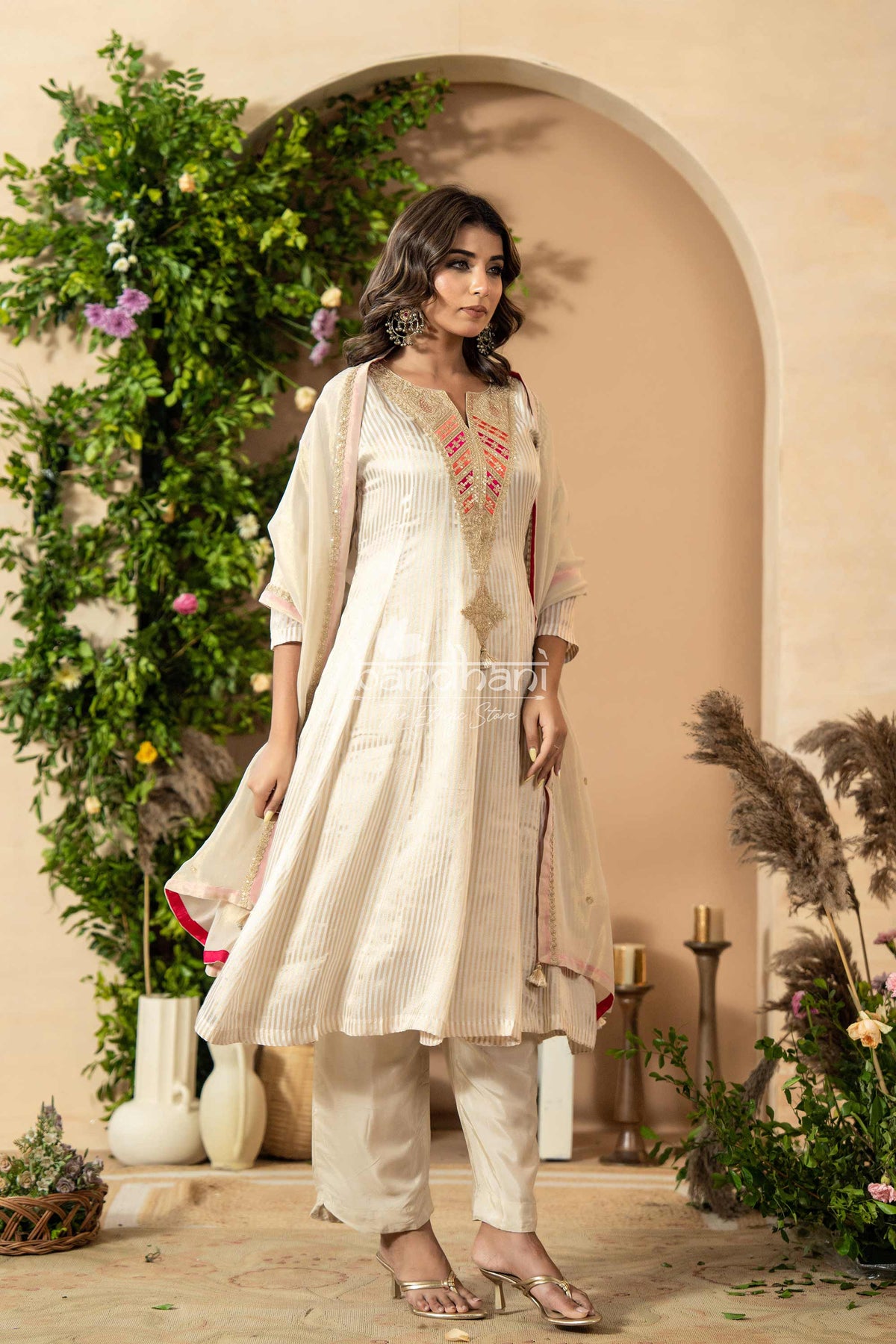 Woman in a white traditional outfit standing in front of a decorative archway with plants.