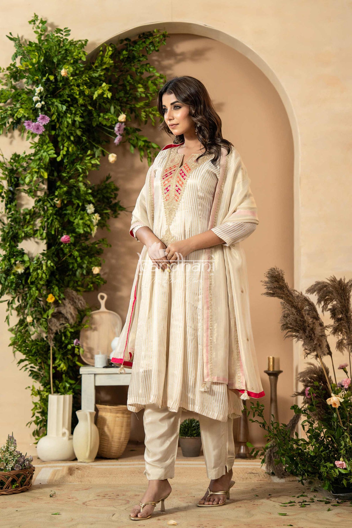 Woman in a traditional outfit standing in a decorated indoor setting with plants and decor.