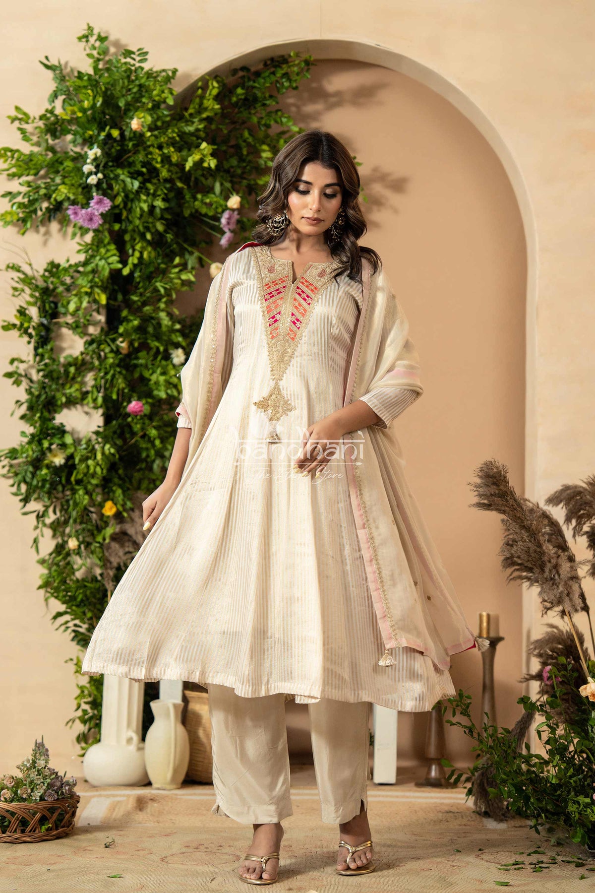 Woman in a traditional outfit standing in a decorative setting with plants and archway.