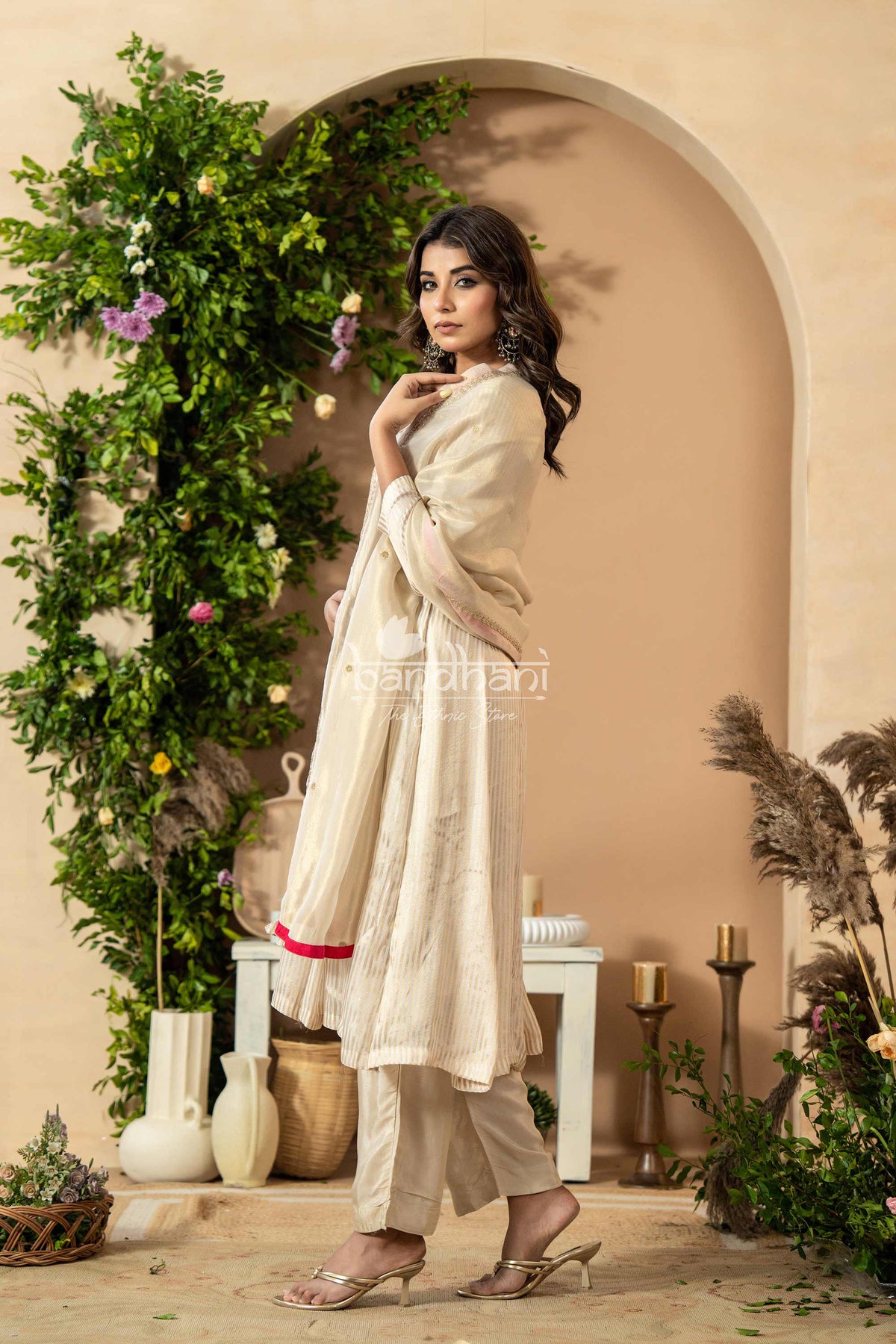 Woman in a white traditional outfit standing in a decorated indoor setting with plants and candles.