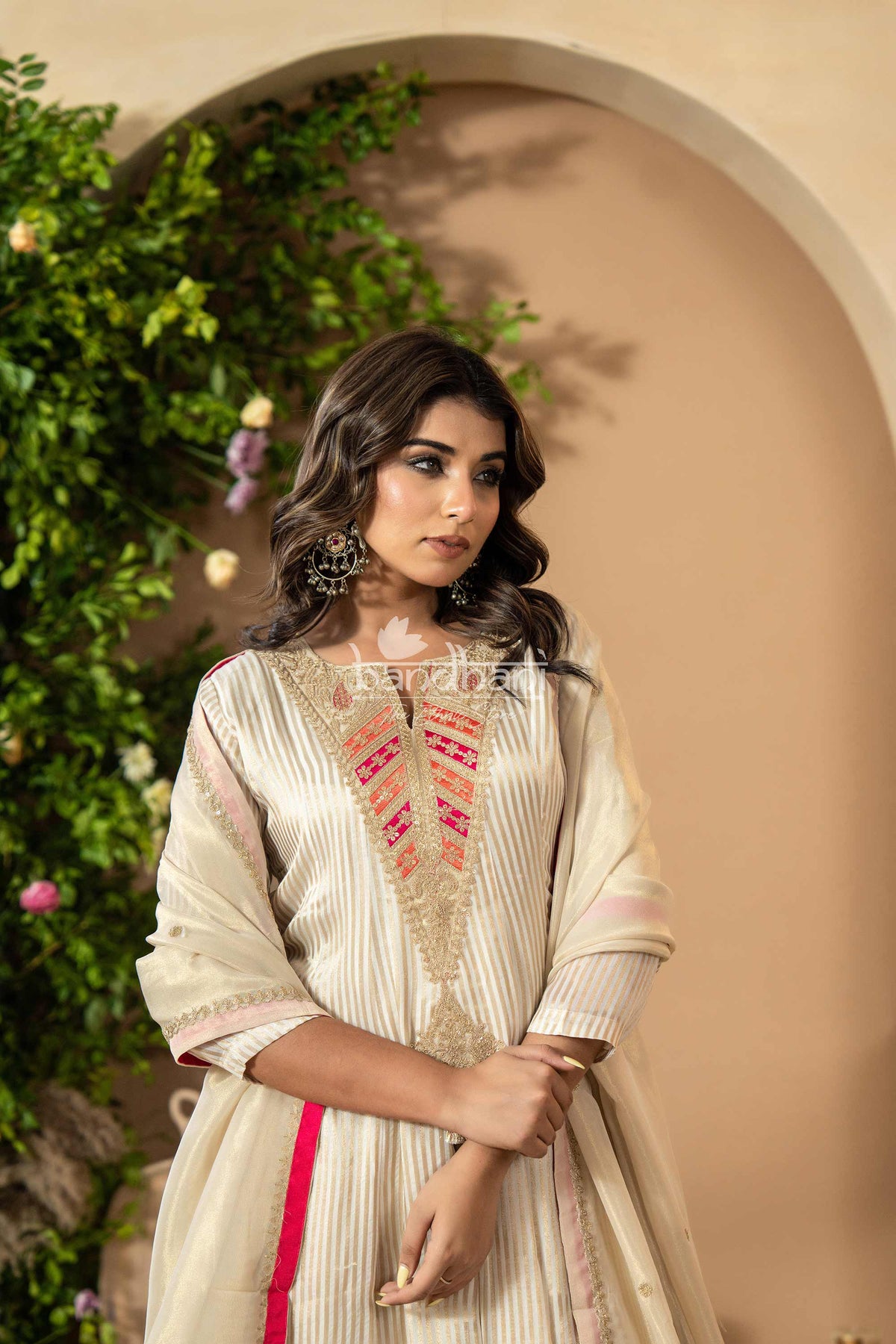 Woman in a traditional outfit with intricate designs against a beige wall and greenery.