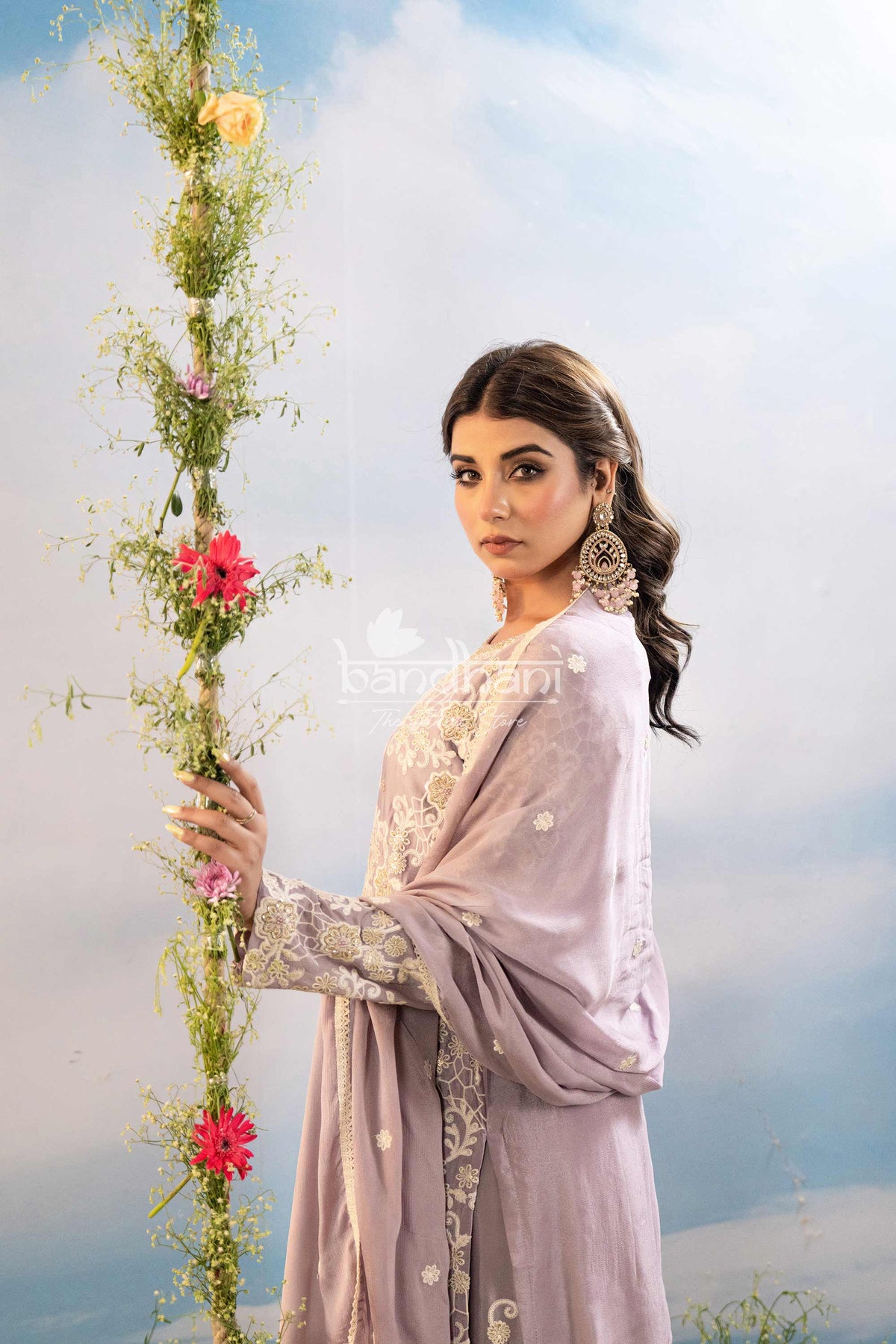 Woman in a light purple saree holding a floral arrangement against a sky background