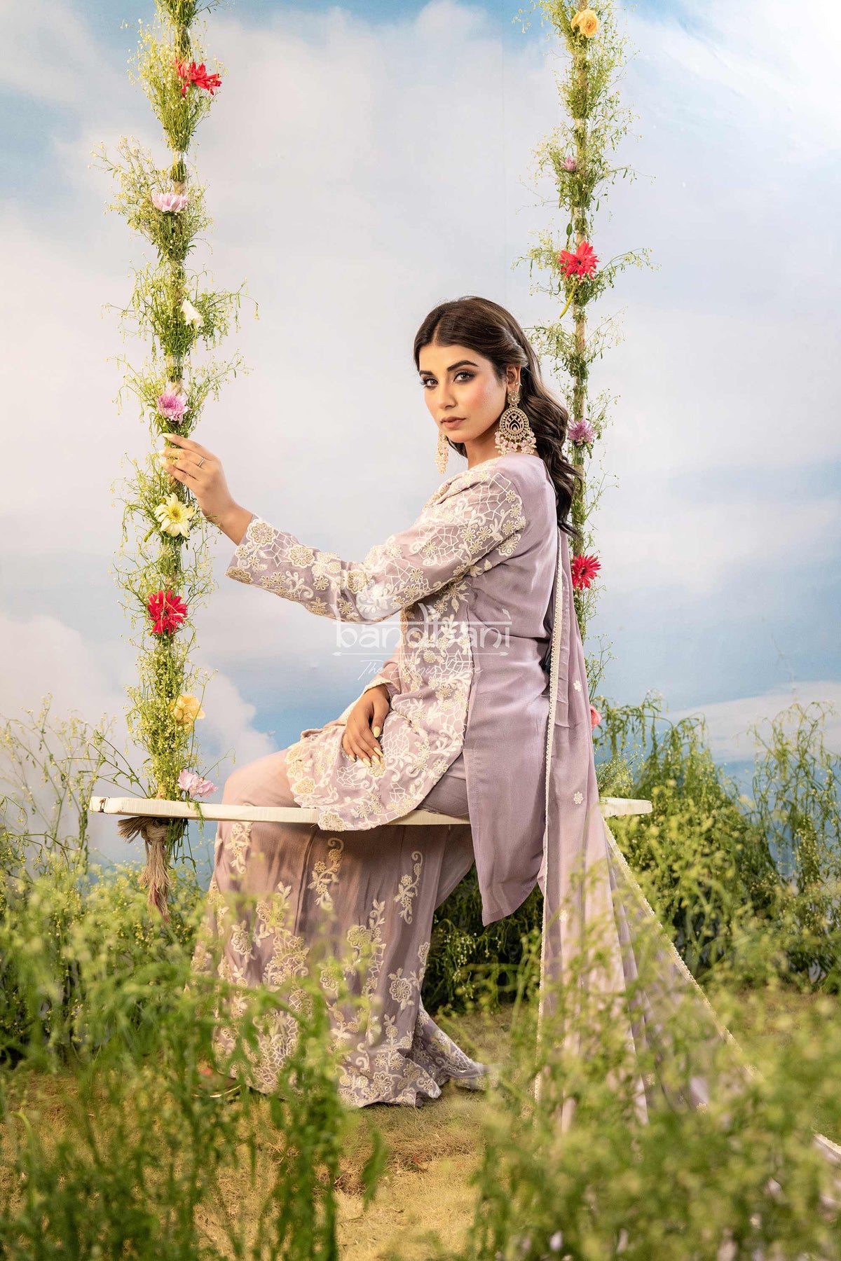 Woman in a light purple traditional outfit sitting on a floral swing with a scenic background.