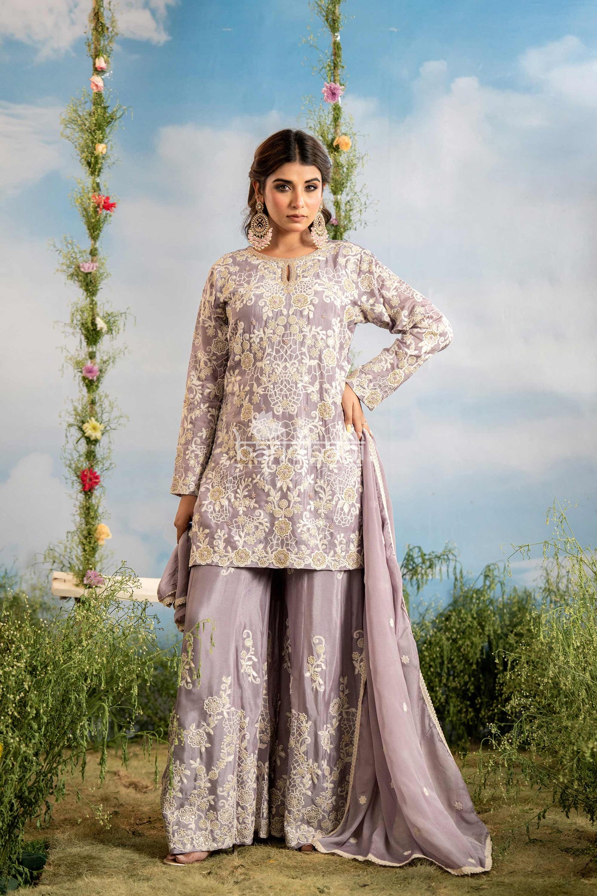 Woman in a traditional outfit standing outdoors with floral decorations and greenery.