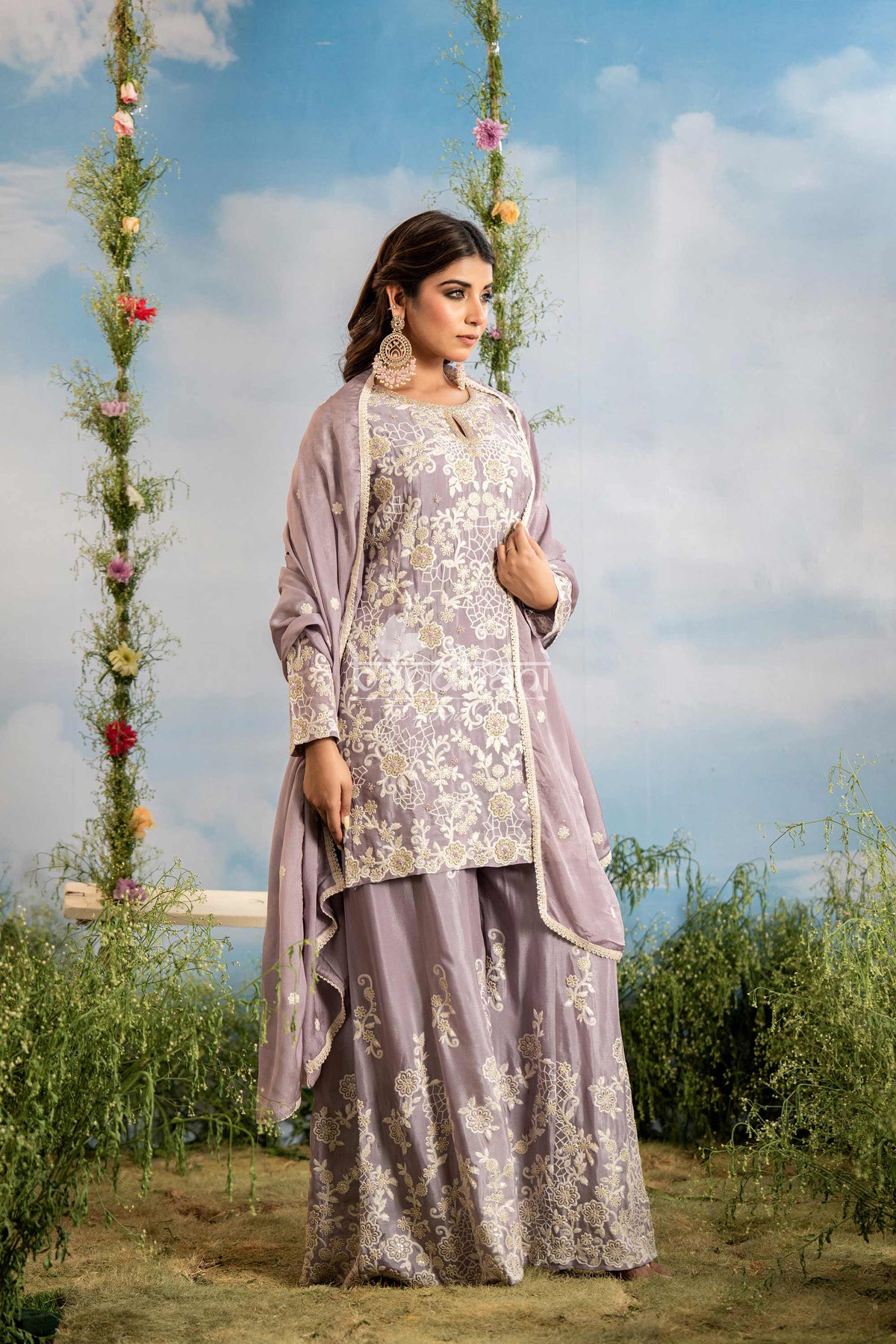 Woman in a lavender traditional outfit with floral patterns standing outdoors against a blue sky.