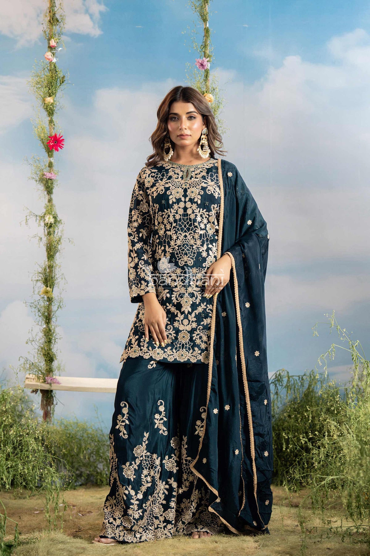 Woman in a blue embroidered traditional outfit standing outdoors with floral decorations.