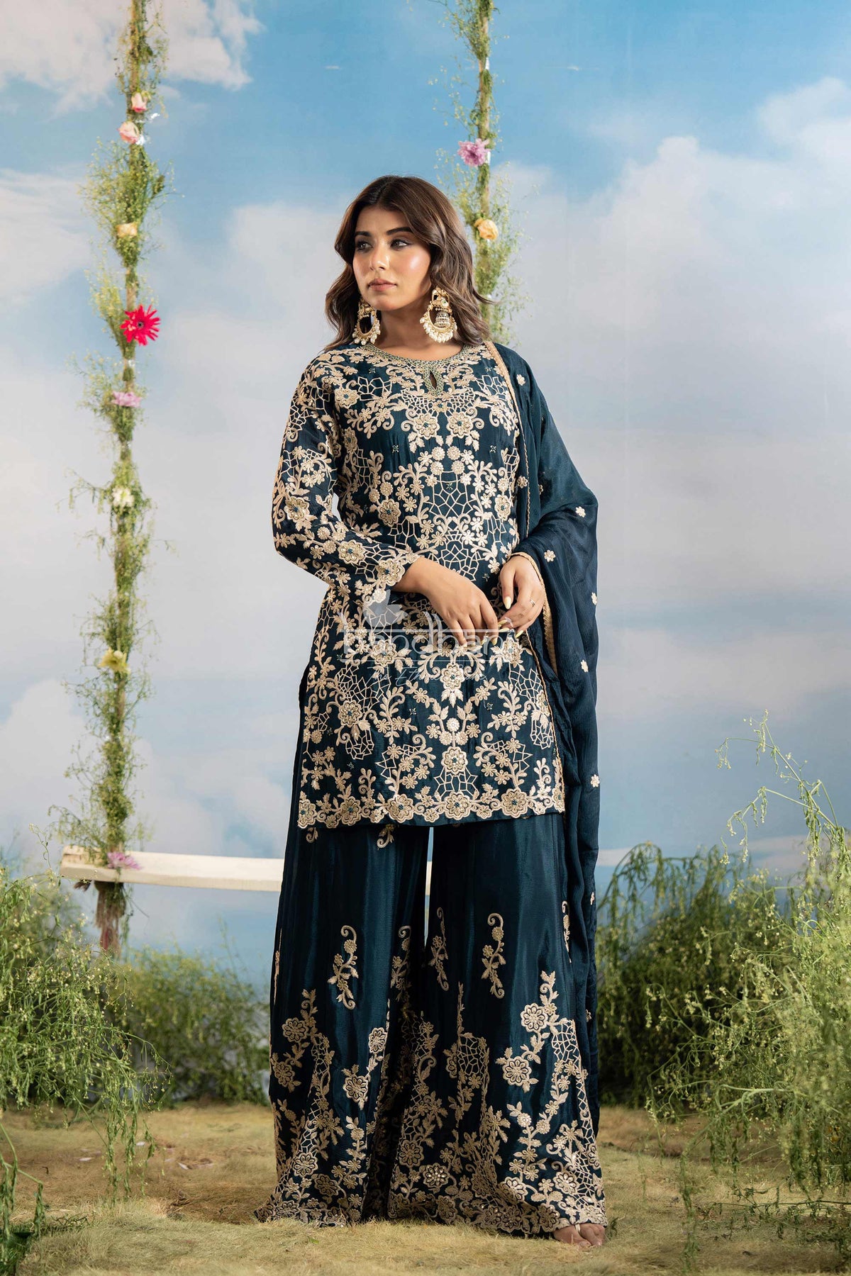 Woman in a floral embroidered outfit standing outdoors with greenery and flowers in the background