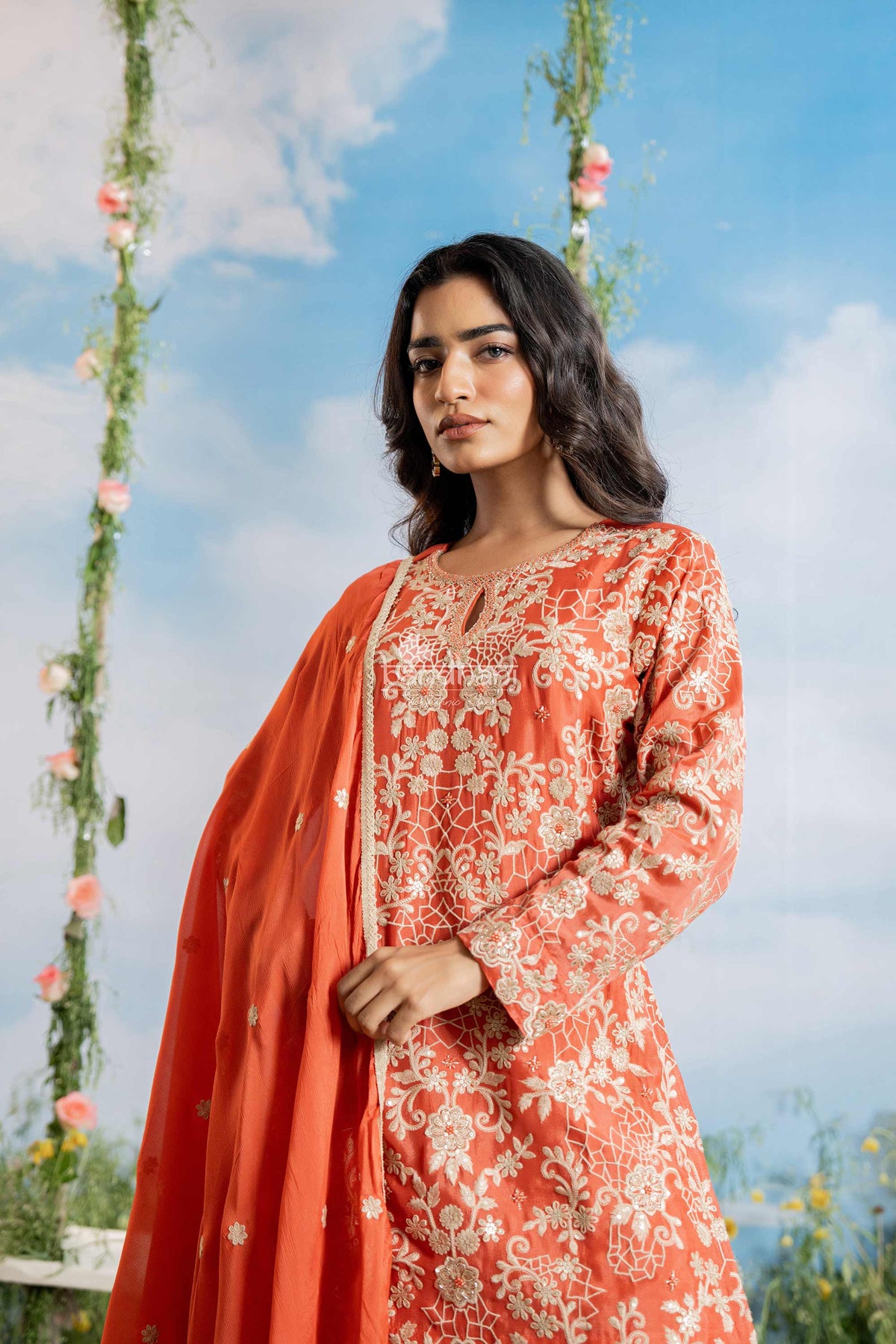Woman in an orange and white embroidered outfit standing outdoors with floral decorations.