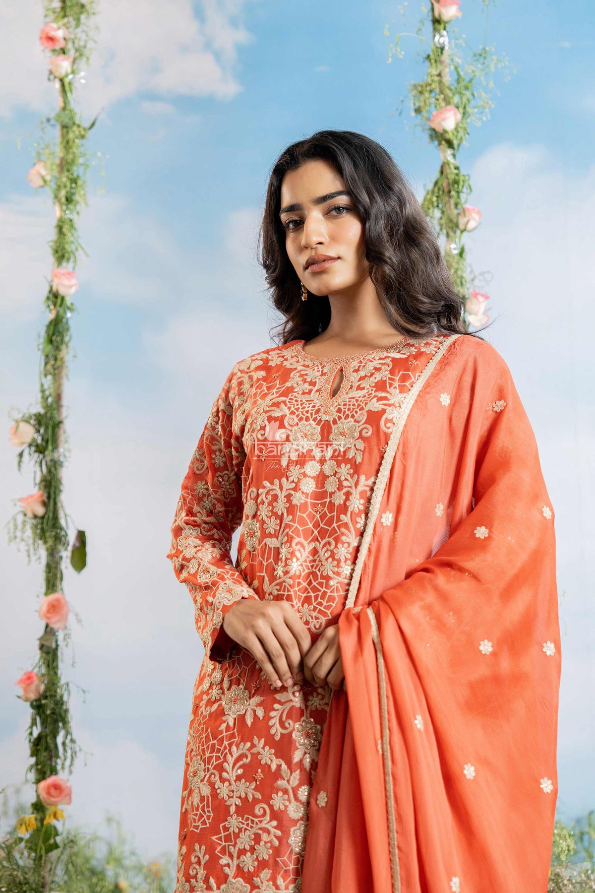 Woman in an orange traditional outfit with floral patterns against a blue sky with flowers.