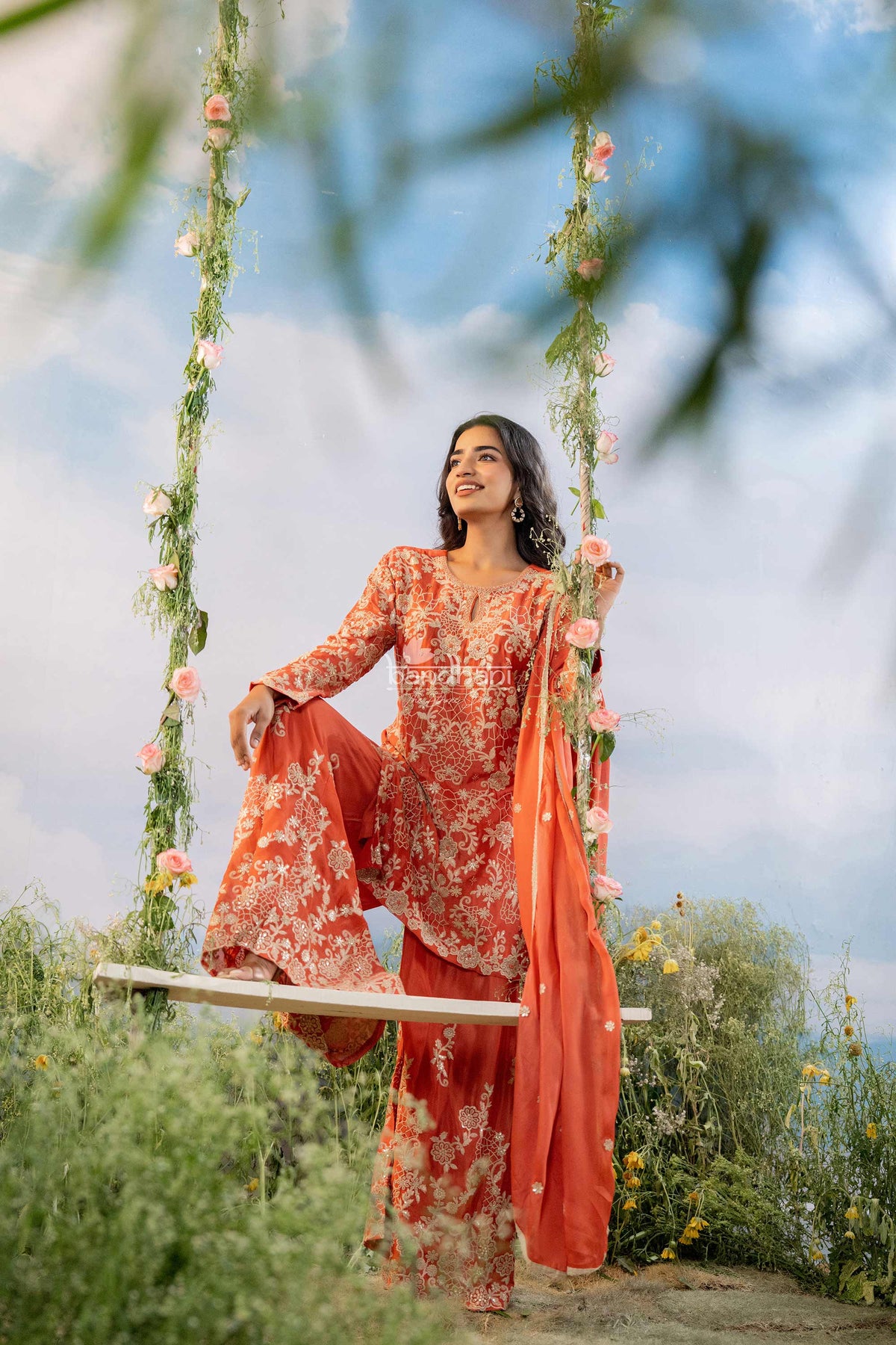 Woman in an orange floral traditional outfit sitting on a swing with a scenic background