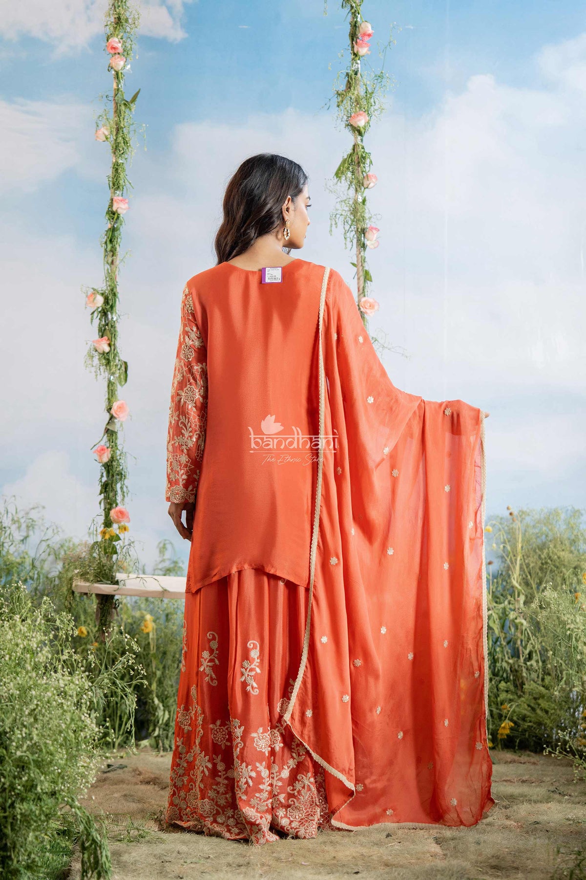 Woman in an orange saree with floral patterns standing on a wooden platform with a blue sky and greenery in the background.