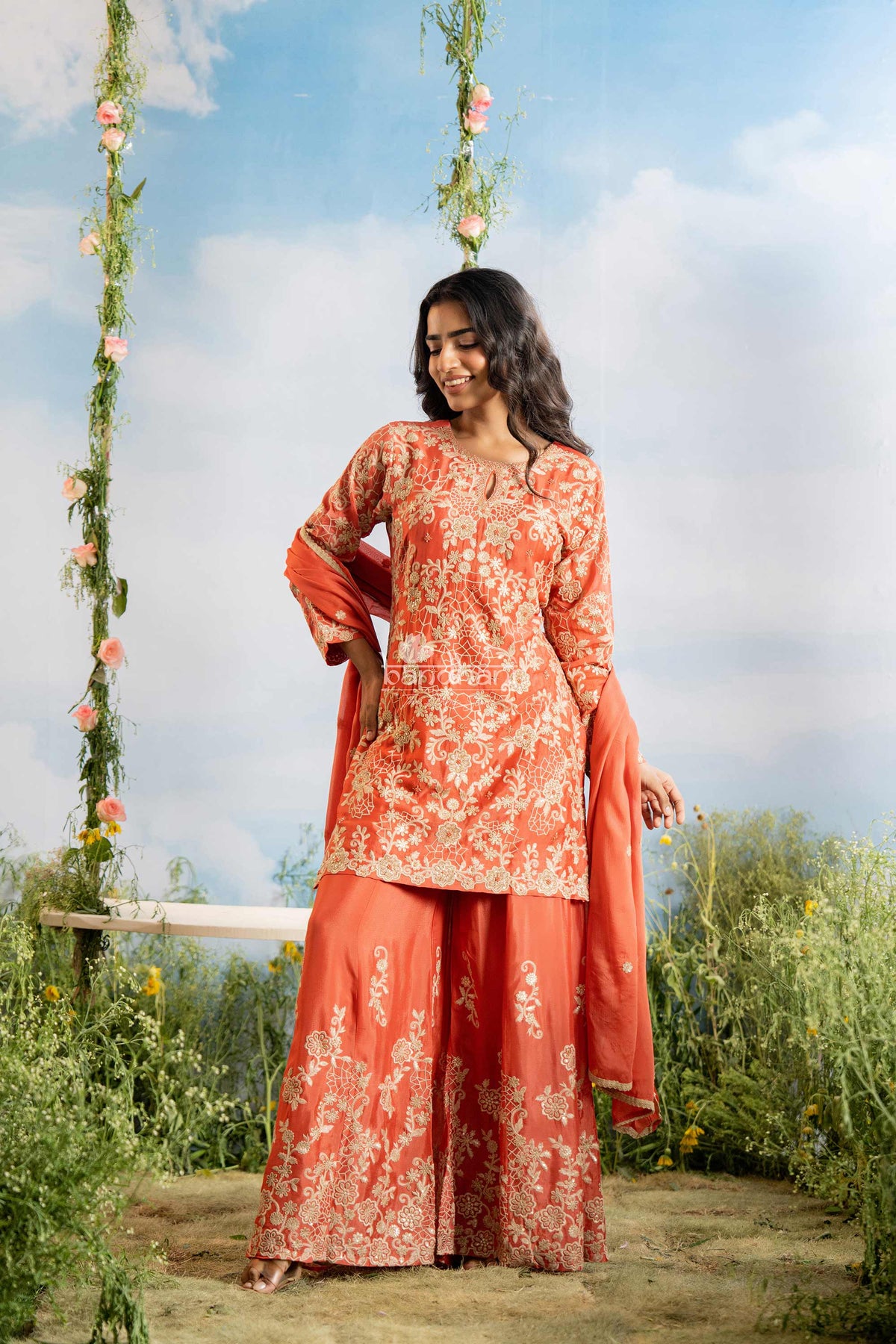 Woman in a red floral dress standing outdoors with greenery and flowers in the background