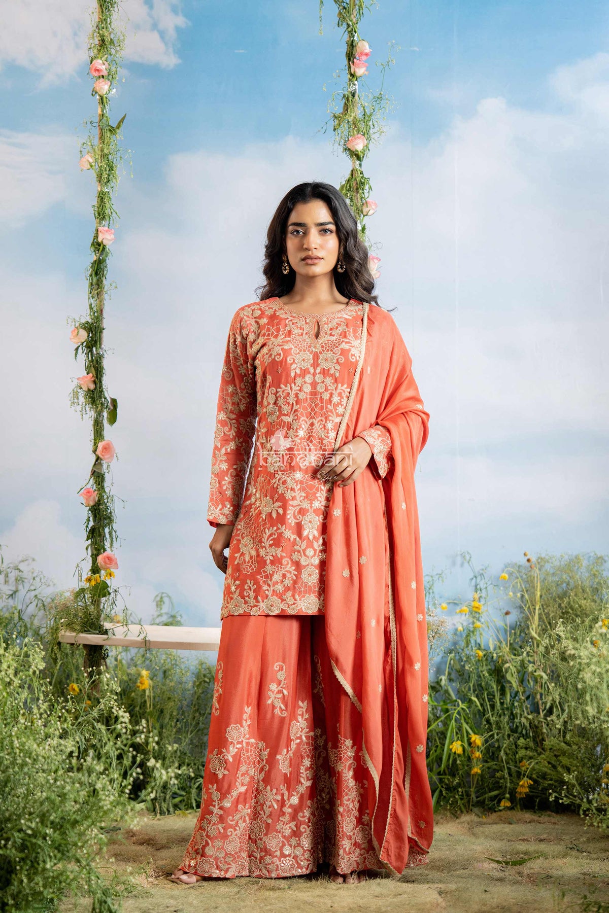 Woman in a coral traditional outfit with floral patterns standing outdoors against a blue sky.